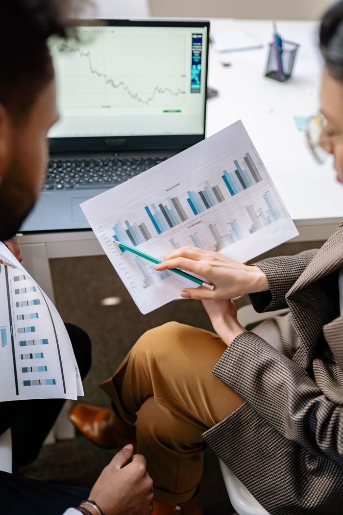 about-img Two adults discuss financial data with charts and laptop in office setting.